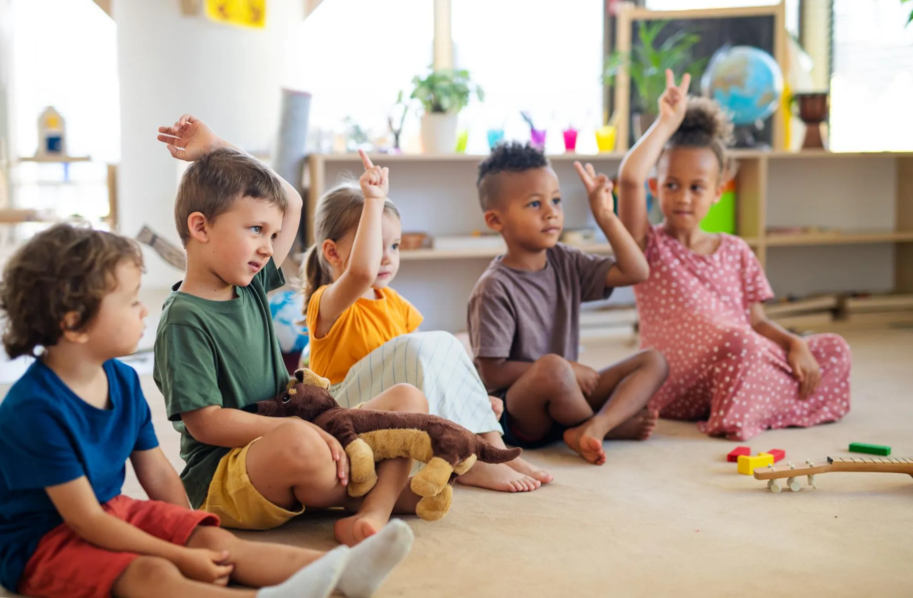 Children raising hands in a preschool setting during a group activity with educational toys and materials around them