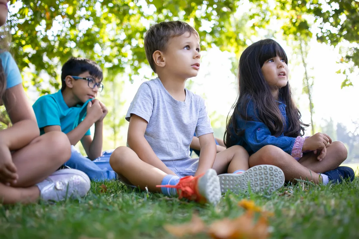 Children sitting on grass outdoors, listening attentively, with trees in the background and sunny weather