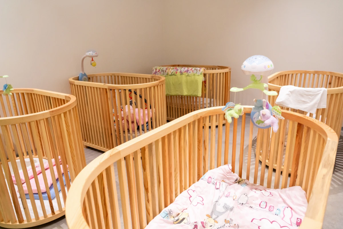 A room with several baby cots arranged, each with soft bedding and colourful toys, providing a safe and nurturing environment for infants.