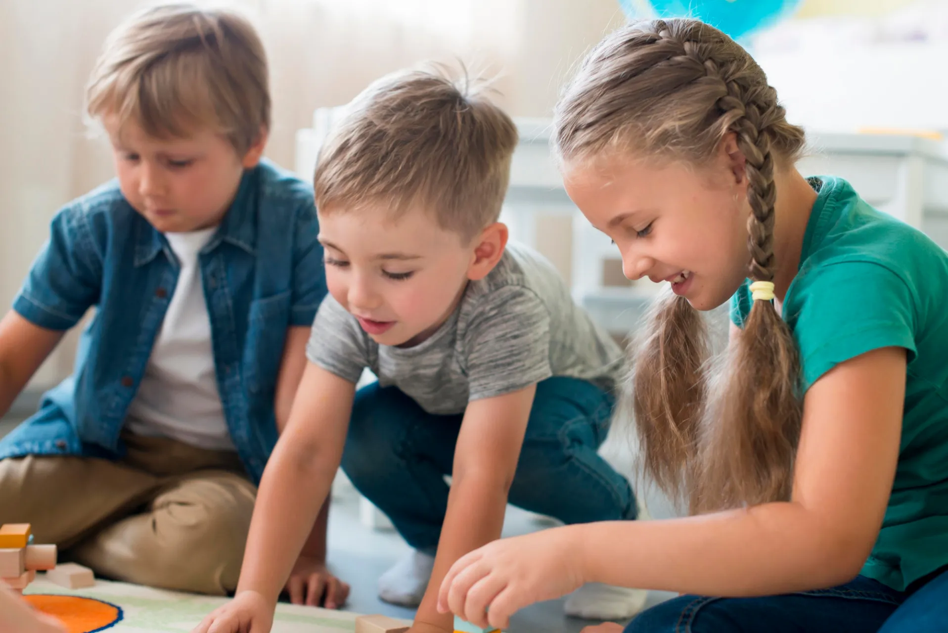 wo boys and a girl playing with building blocks on the floor, engaging in an activity in a bright room.