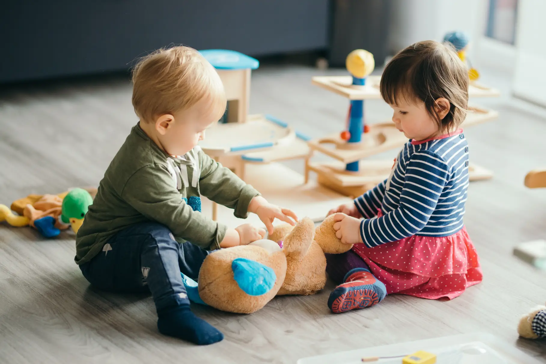 Two toddlers playing with stuffed animals on the floor, exploring and interacting with toys in a warm and safe environment.