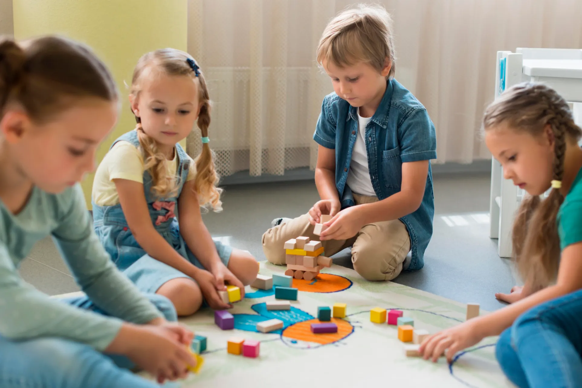 Four children playing with colourful building blocks on a rug, focused on creating structures in a well-lit classroom setting.