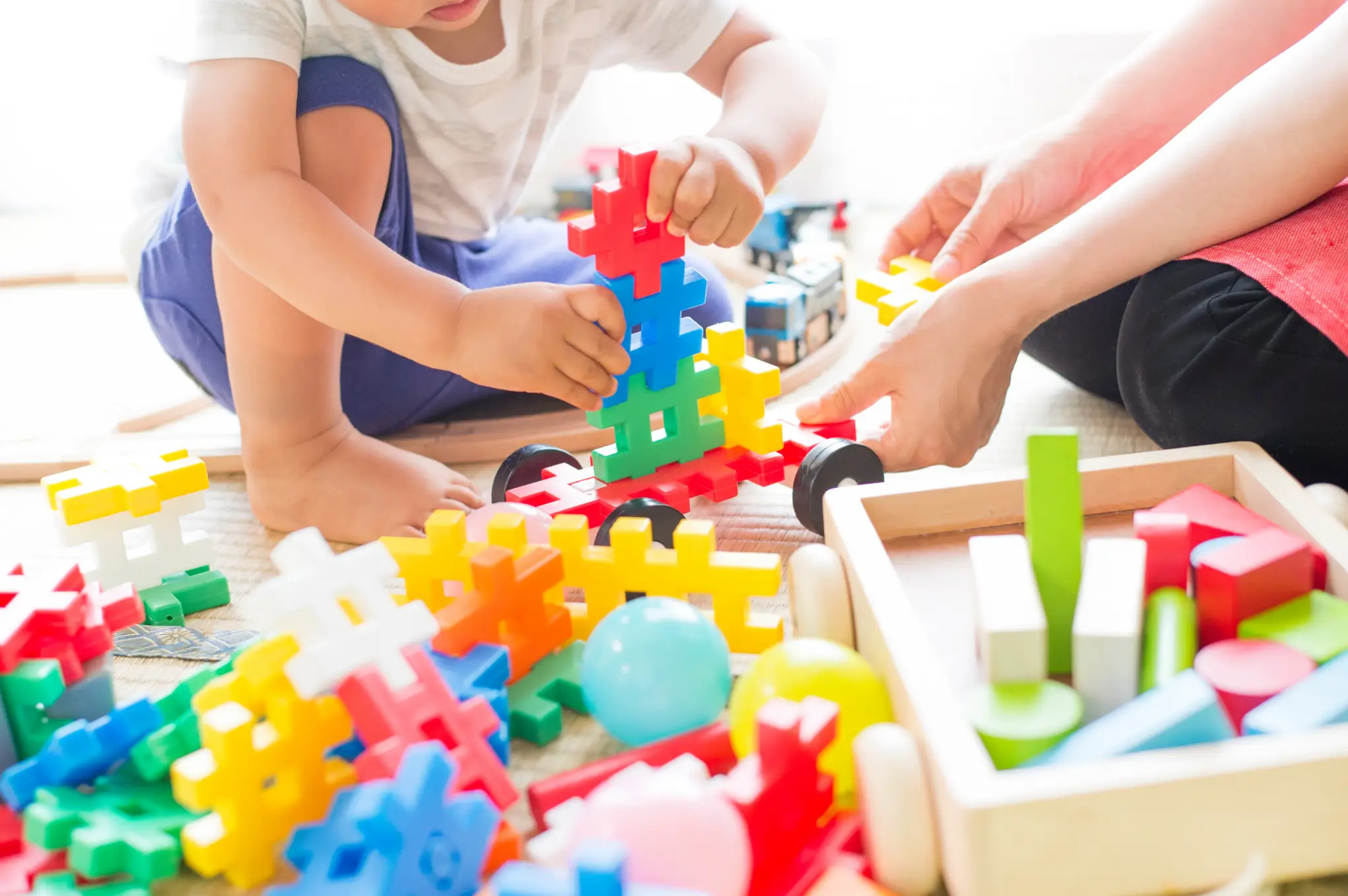 A child focuses on assembling a colourful block structure with assistance from an adult, surrounded by various building toys on the floor.