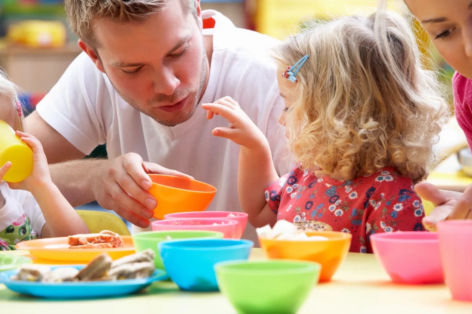 Children and educator sharing a mealtime, helping children develop social skills and independence as they engage with different coloured bowls and cups during lunch.