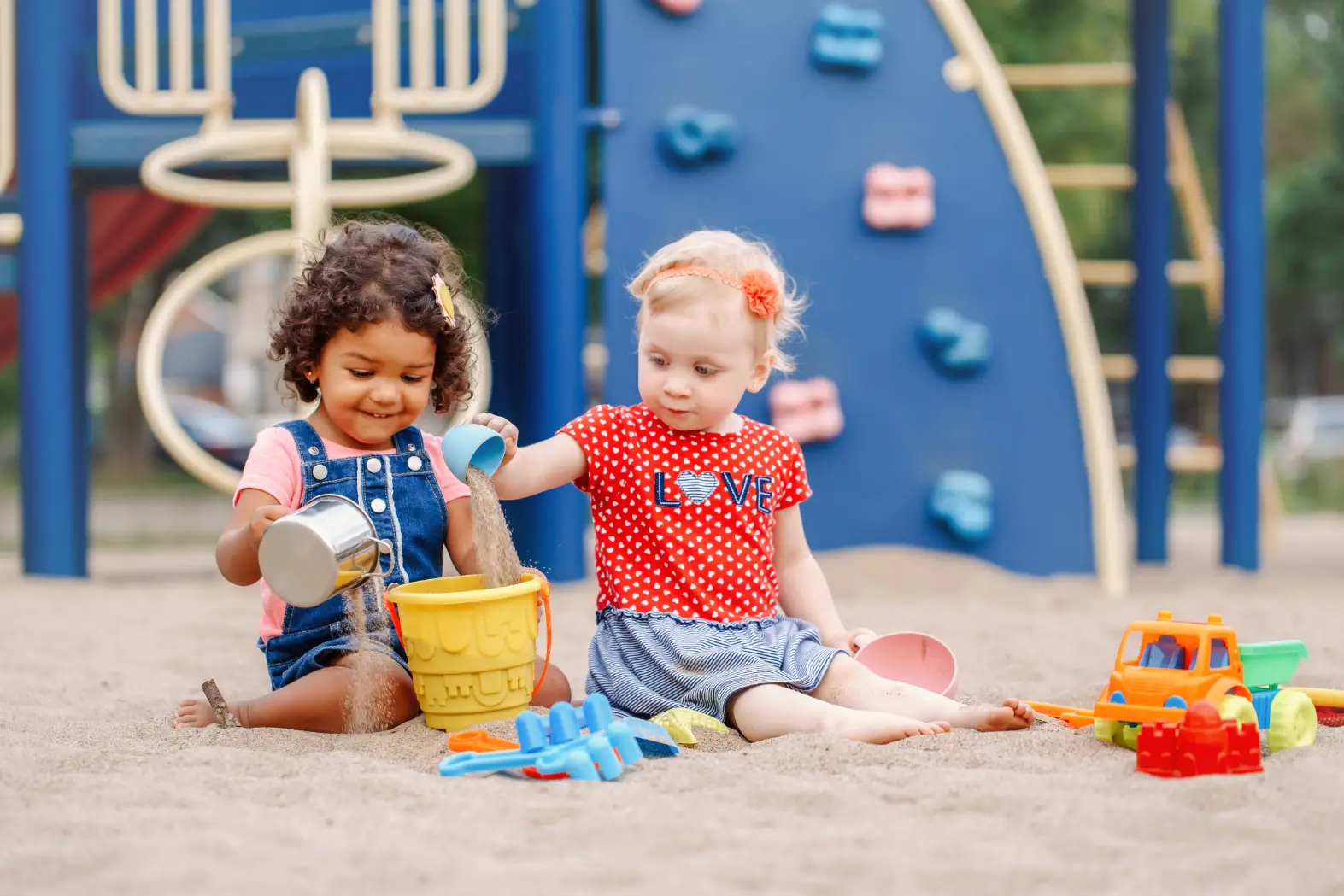 Two young children happily playing with sand toys at a playground, with a bright blue climbing structure in the background.