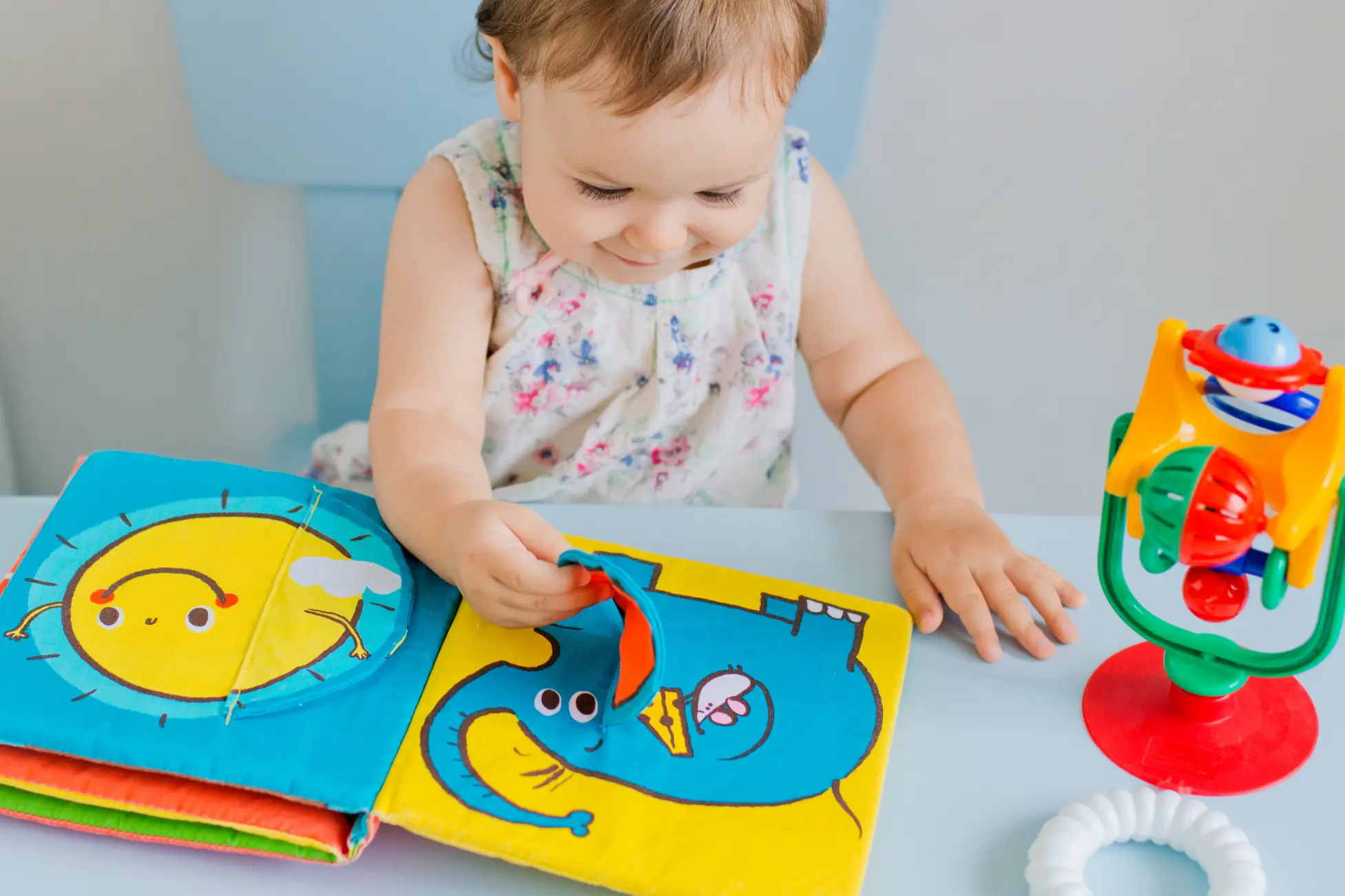 A young child explores a colourful fabric book with animal designs while playing with a toy beside them in a light, welcoming space.