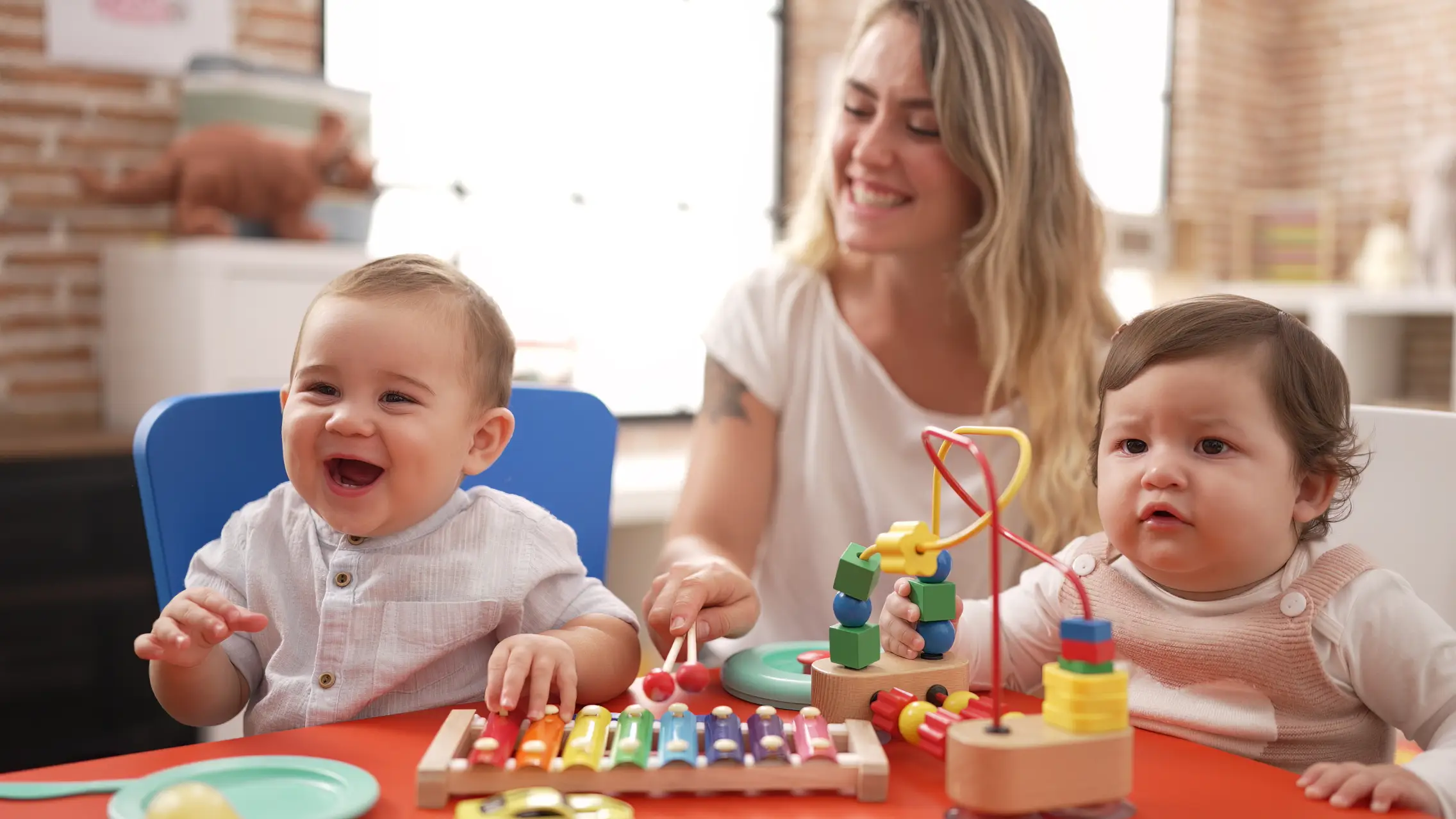 A smiling toddler plays with a colourful wooden xylophone while a baby beside them engages with a toy, both enjoying interactive play with an educator in a bright playroom.