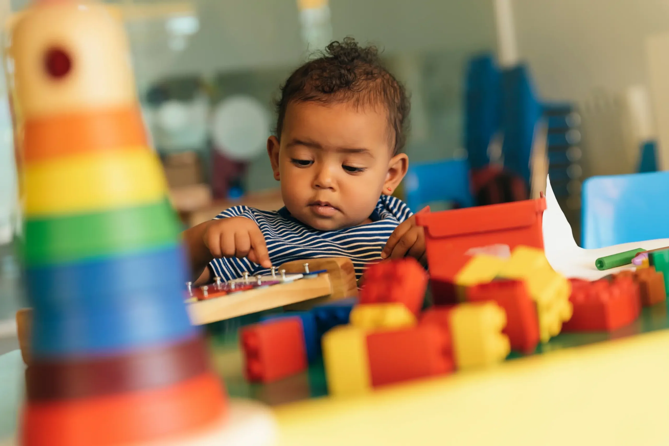 "A baby girl with curly hair holds two colourful balls while sitting with a boy, playing together on a playroom floor with vibrant toys.