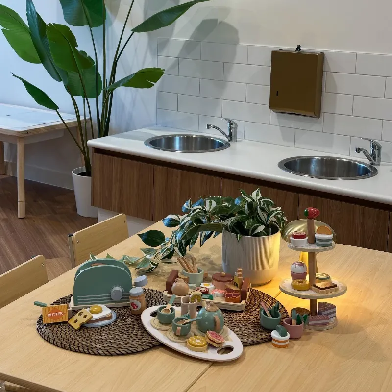 Pretend play setup with wooden kitchen toys and tableware in a modern indoor learning space with double sinks.