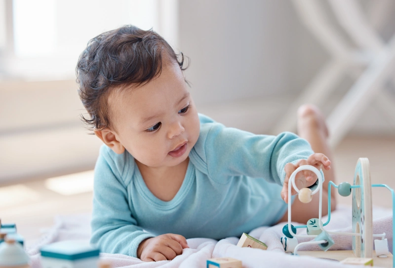A baby focuses on a toy, sitting on a soft mat, with colourful blocks scattered around them, engaged in an interactive learning experience.