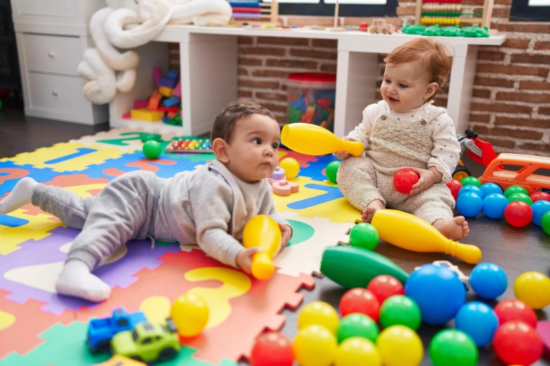 Two babies play with brightly coloured balls and toys on a foam puzzle mat, exploring their surroundings in an early learning environment.