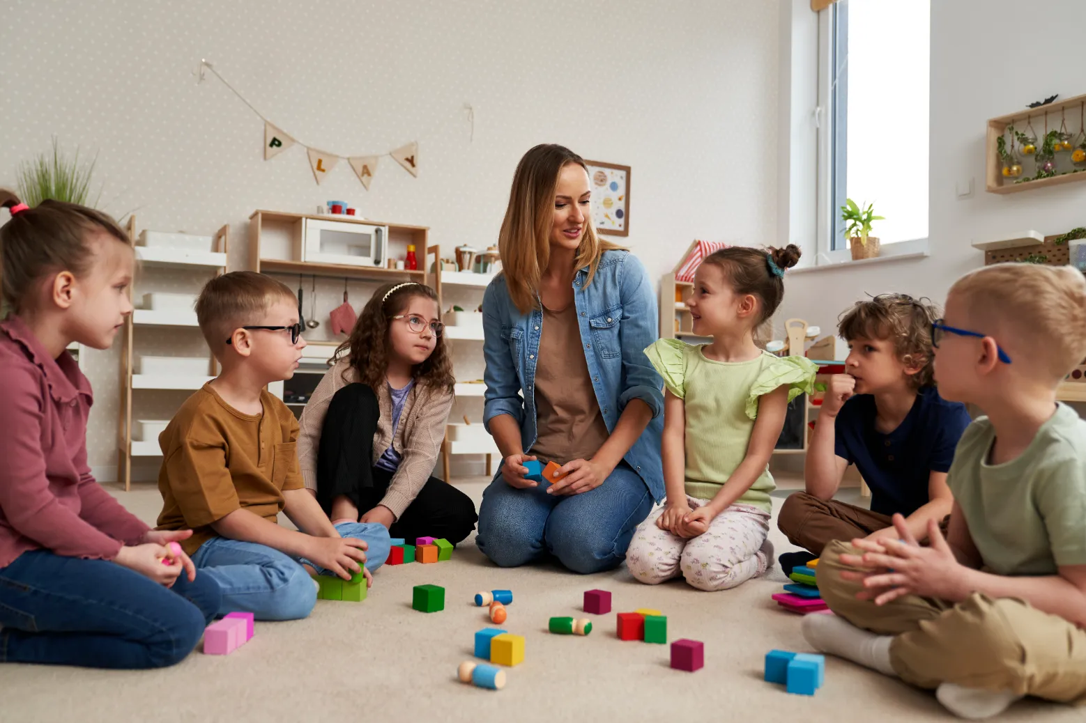 An educator interacting with a group of young children on the floor, engaging in a colourful building block activity in a classroom setting.
