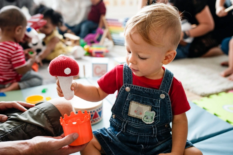 A toddler in a red shirt and denim overalls plays with a toy ice cream cone and building blocks in a lively, colourful playroom.