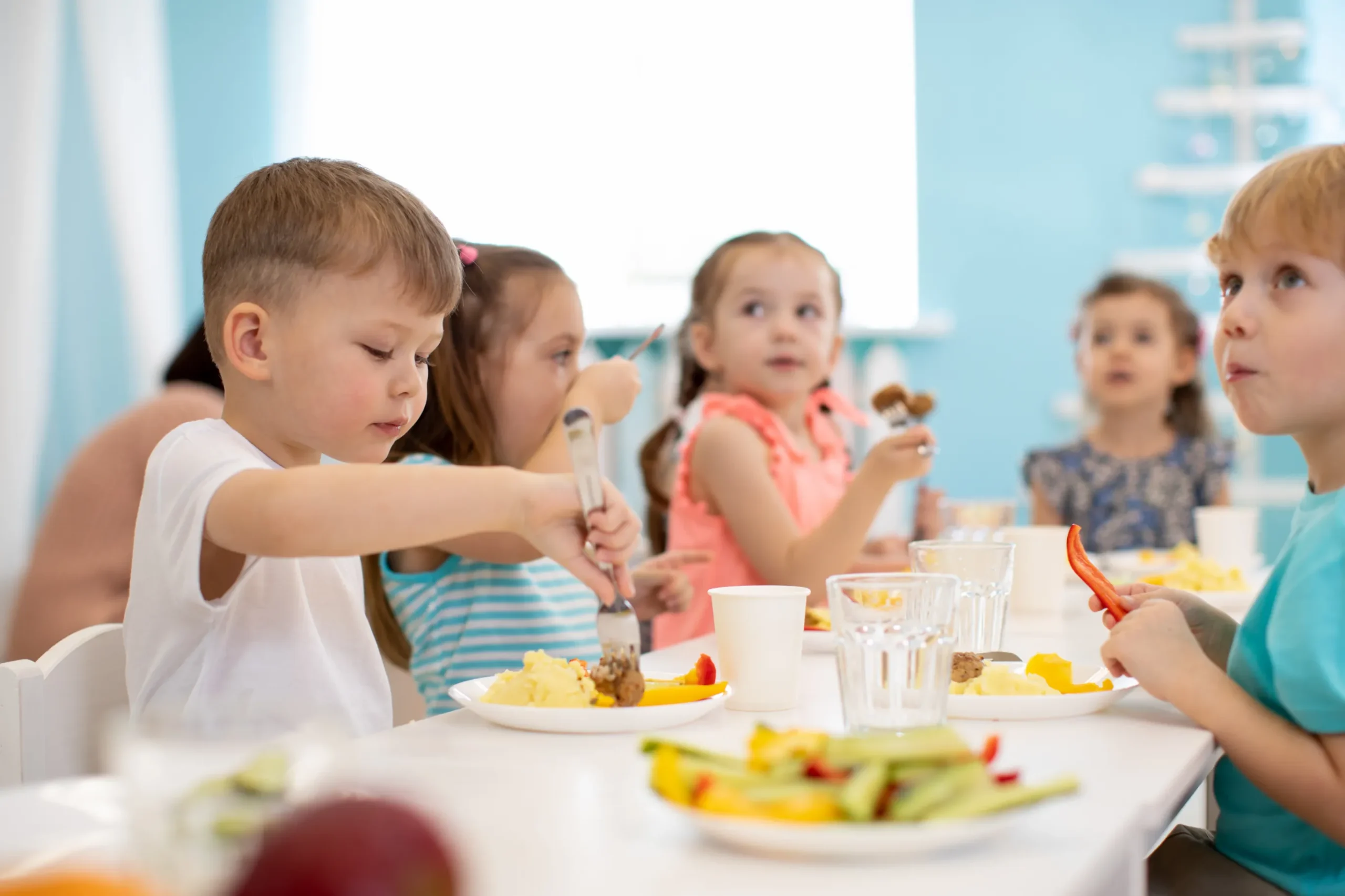 A group of young children eating lunch at a daycare table, with one child eating and others engaging in conversation, surrounded by plates of food and drinks.