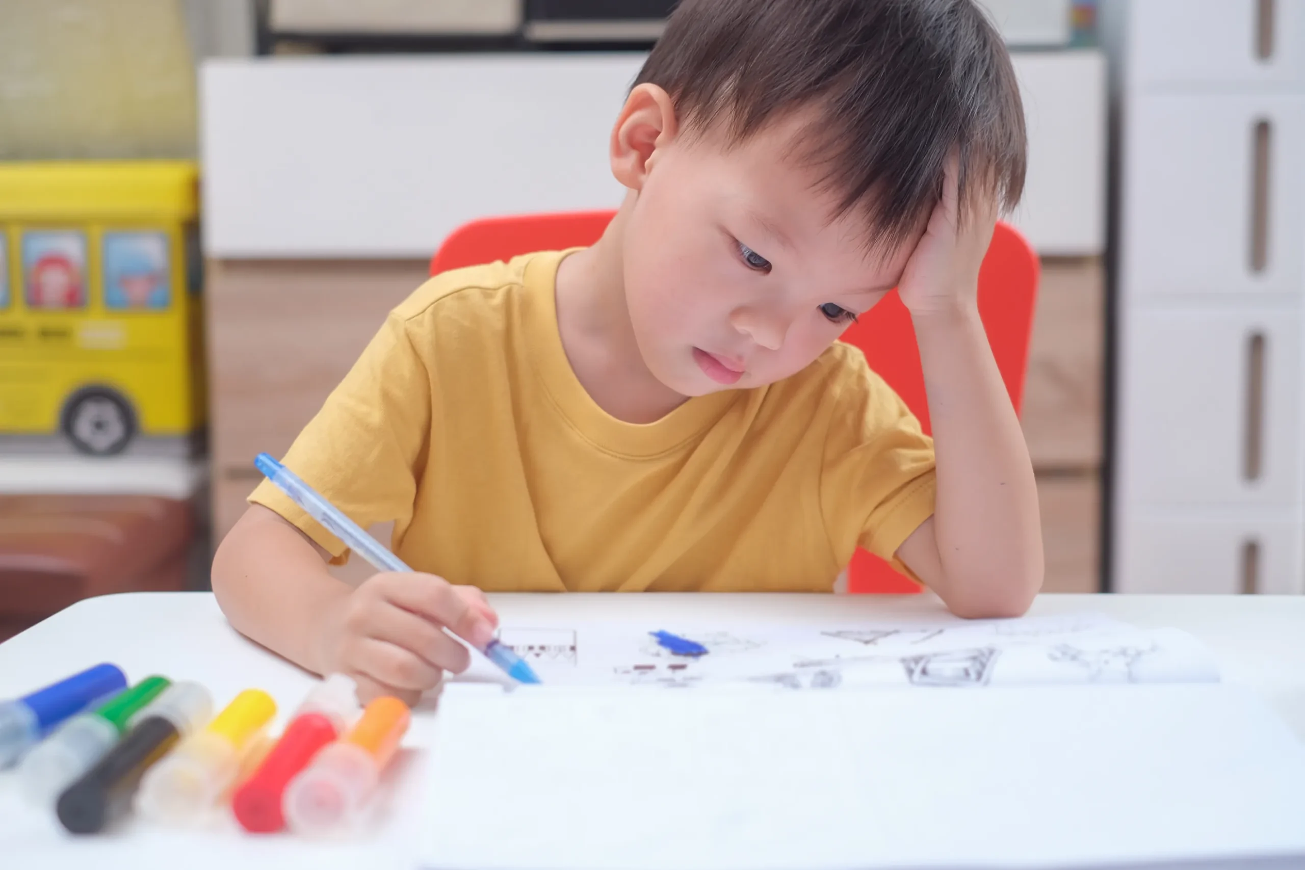 Young boy sitting at a table, concentrating while colouring a picture with markers.