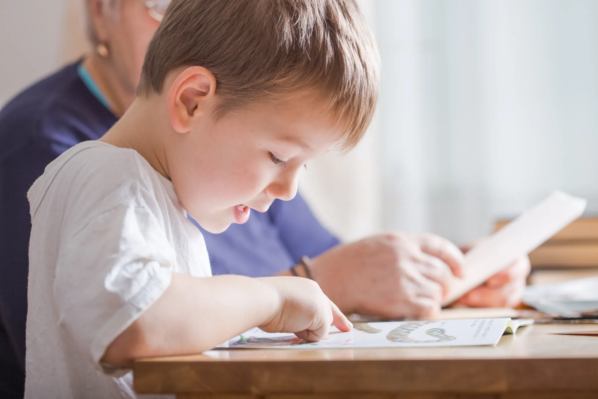 A young boy focused on a book, gently pointing at the page with his right hand while an elderly person is seen in the background.