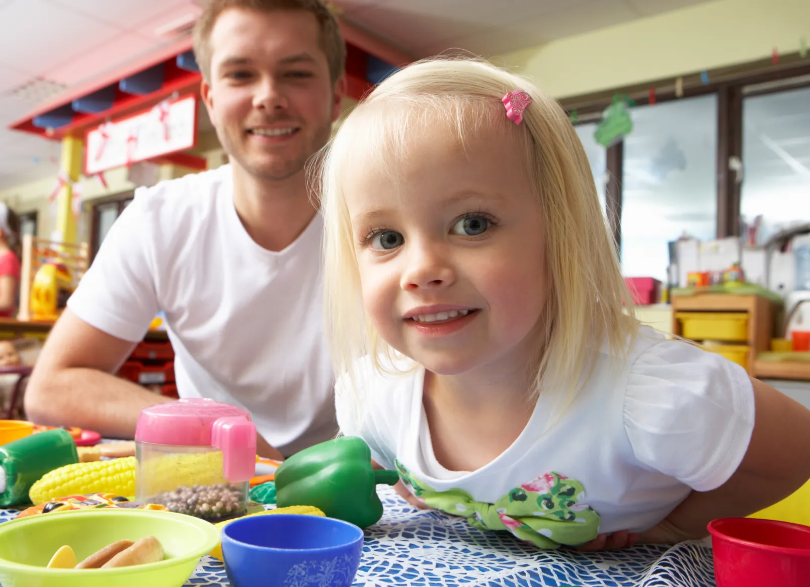 A young girl smiles as she engages in pretend play with toy food and colourful bowls, fostering creativity and social skills in a classroom setting."