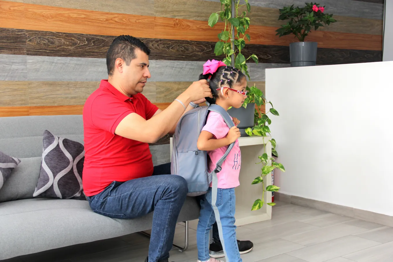 A father helping his daughter put on her backpack, smiling together in a cozy home setting with plants in the background.