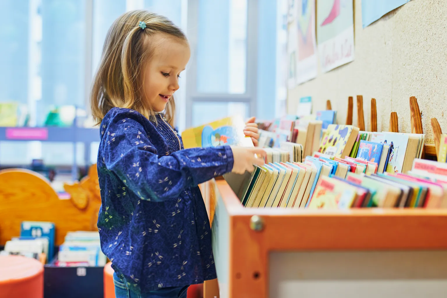 Young girl smiling and selecting a book from a shelf in a classroom setting.